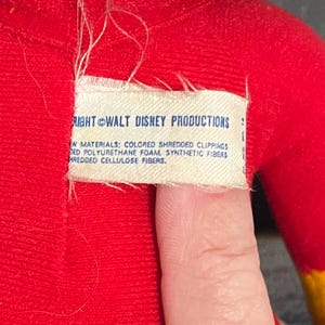 May include: Close-up of a white tag sewn onto a red fabric. The tag reads "RIGHT WALT DISNEY PRODUCTIONS" and lists the materials used in the product: "COLORED SHREDDED CLIPPINGS, DED POLYURETHANE FOAM, SYNTHETIC FIBERS, HREDDED CELLULOSE FIBERS."