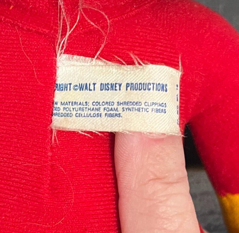 May include: Close-up of a white tag sewn onto a red fabric. The tag reads "RIGHT WALT DISNEY PRODUCTIONS" and lists the materials used in the product: "COLORED SHREDDED CLIPPINGS, DED POLYURETHANE FOAM, SYNTHETIC FIBERS, HREDDED CELLULOSE FIBERS."