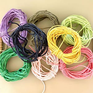 May include: A collection of cordage including seven circular coils of solid-colored string in purple, olive green, black, lime green, yellow, light pink, and dark pink, alongside two small spools of thin twine in yellow-white and red-white patterns.