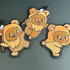 May include: Three embroidered patches featuring brown, furry, bear-like creatures with hoods. Two of the patches depict the creatures holding wooden spears with white wrapped details, while all three display determined facial expressions.