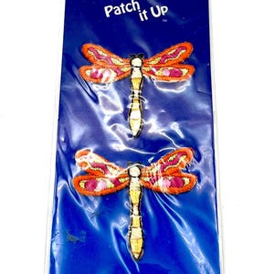 May include: Two dragonfly iron-on patches. Each patch features a dragonfly with orange and pink wings, a yellow and white body, and black accents. The patches are on a blue backing with the text "Patch it Up".