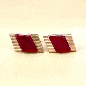 May include: A pair of rectangular cufflinks featuring a central diagonal band of deep red stone set against a metallic silver-toned frame with horizontal grooved detailing on both sides, creating a geometric and structured appearance.
