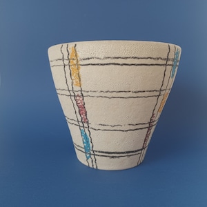 Può includere: Un vaso in ceramica bianca con un motivo geometrico in nero, giallo, blu e rosso. Il vaso ha un'ampia apertura e una base stretta.