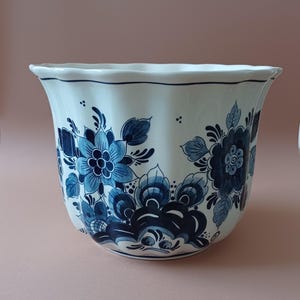Può includere: Un vaso di ceramica bianca con bordo svasato, ornato da un motivo floreale blu. Il design presenta fiori e foglie stilizzate in varie tonalità di blu. Il vaso ha un bordo smerlato e una finitura lucida.