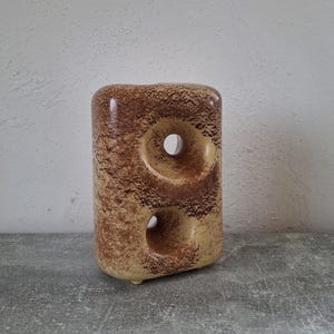 Puede incluir: Objeto decorativo de cerámica con un acabado moteado marrón y beige texturizado. La forma rectangular presenta dos grandes aberturas circulares. La base tiene pequeños pies redondeados. El objeto es probablemente un jarrón o una escultura.