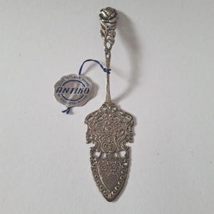 Peut inclure: Une pelle à gâteau antique argentée avec un motif floral. La poignée est ornée d'une rose au sommet, et la pelle présente un motif ajouré orné. Une étiquette avec le mot "ANTIKO" est attachée.