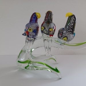 Może przedstawiać: Trzy kolorowe szklane ptaki siedzące na przezroczystej szklanej gałęzi. Ptaki są wykonane ze szkła Murano i mają skomplikowane detale. Gałąź jest zielona i ma spiralny wzór.