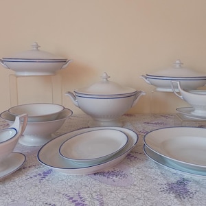 Peut inclure: Un ensemble de plats de service en porcelaine blanche avec une bordure bleue. L'ensemble comprend un grand bol de service, deux bols plus petits, deux saucières et deux assiettes. Les plats sont disposés sur une nappe blanche.