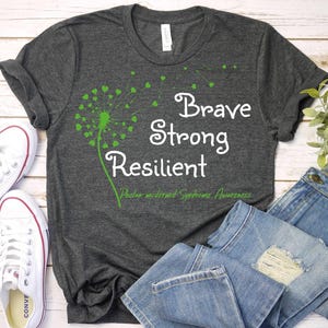 May include: A dark gray t-shirt with a green dandelion design and the words "Brave Strong Resilient" in white. The text "Phelan-McDermid Syndrome Awareness" is also included in white.