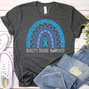 May include: A dark grey t-shirt with a blue rainbow design and a blue ribbon in the center. The text "Behçet's Disease Awareness" is printed below the rainbow.
