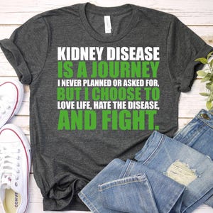 May include: Dark gray t-shirt with the text "KIDNEY DISEASE IS A JOURNEY" in white and green. The shirt also includes the phrases "I NEVER PLANNED OR ASKED FOR, BUT I CHOOSE TO LOVE LIFE, HATE THE DISEASE, AND FIGHT."