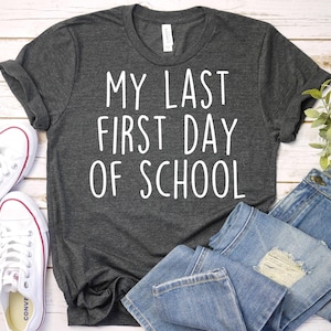 May include: Dark gray t-shirt with the text "MY LAST FIRST DAY OF SCHOOL" in white. The shirt is paired with blue jeans and white sneakers. A casual outfit for back-to-school or graduation.