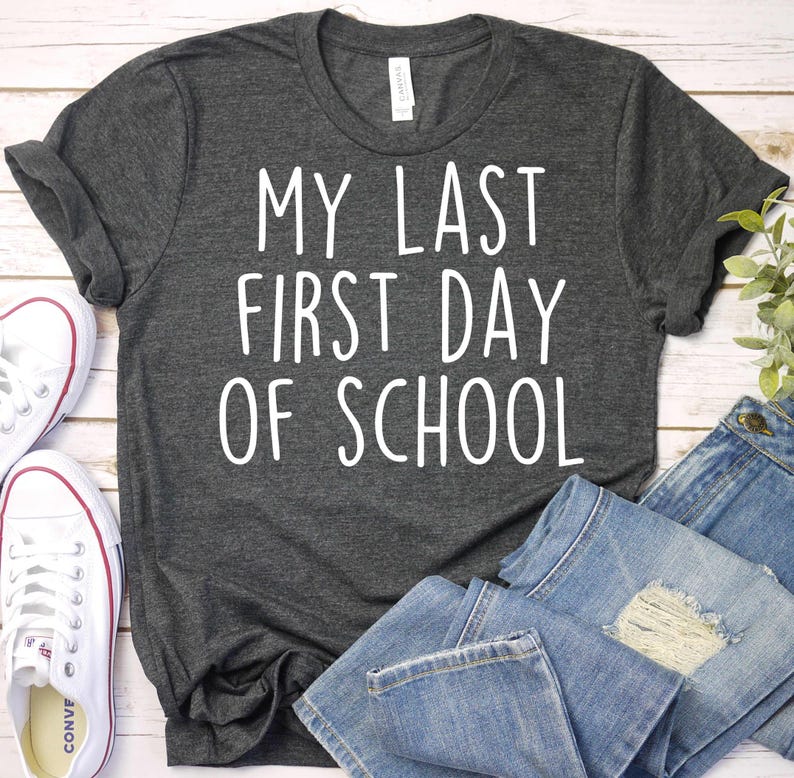 May include: Dark gray t-shirt with the text "MY LAST FIRST DAY OF SCHOOL" in white. The shirt is paired with blue jeans and white sneakers. A casual outfit for back-to-school or graduation.