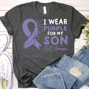 May include: A dark gray t-shirt with a purple ribbon, a feather, and the text "I wear purple for my son CDLS Awareness".