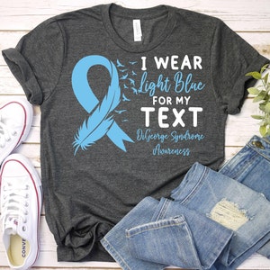 May include: A dark gray t-shirt with a light blue ribbon and a feather design. The text on the shirt reads "I wear light blue for my text DiGeorge Syndrome Awareness".