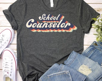 May include: A dark gray t-shirt with a retro-style graphic that says "School Counselor" in colorful, layered letters.
