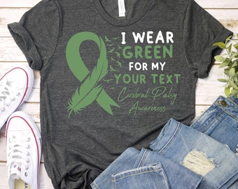 May include: A dark gray t-shirt with a green ribbon and feather design. The text on the shirt reads "I wear green for my [your text] Cerebral Palsy Awareness."