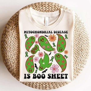 May include: Cream-colored t-shirt with the text "MITOCHONDRIAL DISEASE IS BOO SHEET." The shirt features green ghost illustrations with floral and bat designs. Green awareness ribbons and flowers are also included in the design.