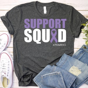 May include: A dark gray t-shirt with the text "SUPPORT SQUAD" in white with a purple ribbon. The text "#SMASAWARENESS" is below the ribbon.