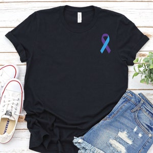May include: Black t-shirt with a purple and blue ribbon on the left chest. The t-shirt is laying on a white wooden surface with a pair of white sneakers and blue denim shorts.