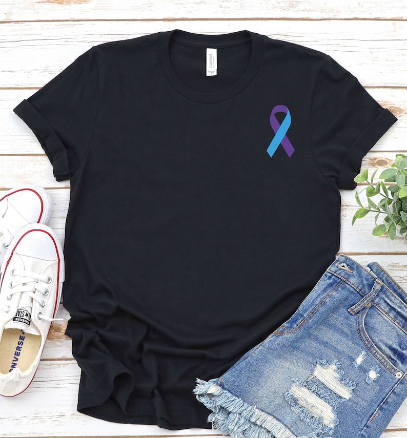 May include: Black t-shirt with a purple and blue ribbon on the left chest. The t-shirt is laying on a white wooden surface with a pair of white sneakers and blue denim shorts.