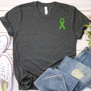 May include: A dark heather grey t-shirt with a green ribbon on the front. The shirt is laid flat on a white wooden surface with a pair of white sneakers and blue jeans.