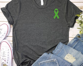 Puede incluir: Una camiseta gris oscuro jaspeada con una cinta verde en la parte delantera. La camiseta está colocada sobre una superficie de madera blanca con un par de zapatillas blancas y unos vaqueros azules.