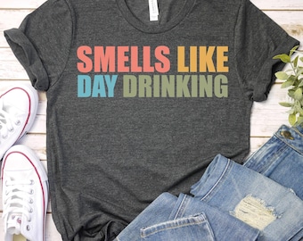 Puede incluir: Camiseta gris oscuro con la frase "Smells Like Day Drinking" en letras coloridas. Se combina con vaqueros azules y zapatillas blancas, lo que sugiere un estilo casual y relajado. La camiseta está hecha de un material suave.