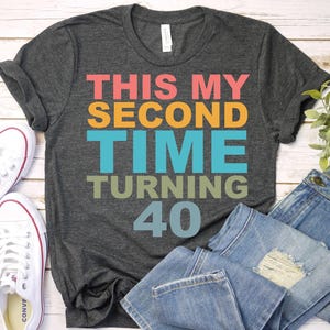 May include: Dark gray t-shirt with the text "THIS MY SECOND TIME TURNING 40" in pink, orange, blue, and green. White canvas sneakers and blue jeans are also in the image. The shirt is a casual style.