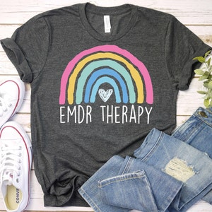 K&ouml;nnte beinhalten: Ein dunkelgraues T-Shirt mit einem Regenbogen- und einem Herzmotiv. Der Text "EMDR THERAPY" ist unter dem Regenbogen aufgedruckt.