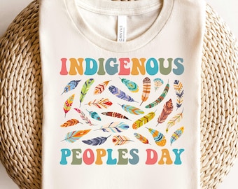 May include: A beige t-shirt with the text "Indigenous Peoples Day" in colorful letters. The text is surrounded by a pattern of colorful feathers.