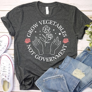 K&ouml;nnte beinhalten: Ein dunkelgraues T-Shirt mit einem wei&szlig;en Grafikdesign, auf dem "Grow Vegetables Not Government" mit einer Blume und zwei H&auml;nden, die sie halten, steht. Die Grafik befindet sich in einem Kreis mit einer roten Rose auf jeder Seite.
