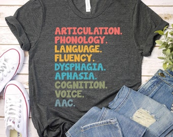 May include: A dark grey t-shirt with a colourful graphic design listing the terms: Articulation, Phonology, Language, Fluency, Dysphagia, Aphasia, Cognition, Voice, AAC.