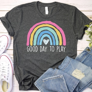 May include: Dark gray t-shirt with a colorful rainbow design and the text "GOOD DAY TO PLAY". The rainbow features pink, yellow, teal, and blue arches with a white heart in the center. White sneakers and blue jeans are also in the image.