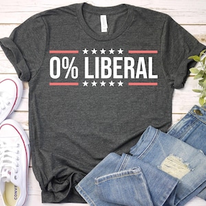 May include: Dark gray t-shirt with the text "0% LIBERAL" in white, framed by red lines and stars. The shirt is paired with blue jeans and white sneakers.