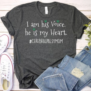 May include: A dark gray t-shirt with the text "I am his Voice. he is my Heart. #CerebralPalsyMom" printed in white. The shirt is laying on a wooden surface with a pair of white sneakers and blue jeans.