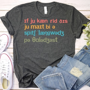 May include: Dark gray t-shirt with the text "If ju kæn rid ðis ju maıt bi ə spits længwadz pə ðalǝdzəst" in various colors. The shirt is paired with blue jeans and white canvas sneakers.