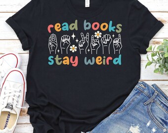 May include: Black t-shirt with the text "read books stay weird" in colourful letters. The text is accompanied by a row of hand gestures in sign language.