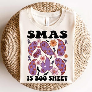 May include: A white t-shirt with a black graphic that says "SMAS IS BOO SHEET" with purple ghosts, bats, and flowers.