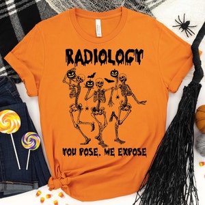 May include: Orange t-shirt with the word "RADIOLOGY" above a graphic of dancing skeletons holding jack-o'-lanterns. The text "YOU POSE. ME EXPOSE" is below the skeletons. Halloween-themed apparel.