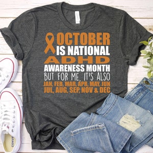 May include: A dark gray t-shirt with an orange ribbon and the text "OCTOBER IS NATIONAL ADHD AWARENESS MONTH BUT FOR ME IT'S ALSO JAN, FEB, MAR, APR, MAY, JUN JUL, AUG, SEP, NOV & DEC"