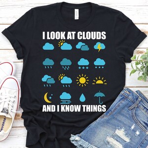 May include: Black t-shirt with the text "I look at clouds and I know things" and a variety of weather icons in blue, yellow, and white.
