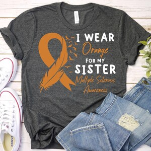May include: Gray t-shirt with an orange ribbon and feather design. The text reads "I wear orange for my sister Multiple Sclerosis Awareness."