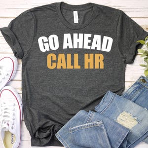 May include: Dark gray t-shirt with the text "GO AHEAD CALL HR" in white and orange. The shirt is paired with white sneakers and blue jeans.