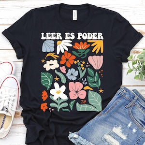 May include: Black t-shirt with a colorful floral design and the text "LEER ES PODER" in white. The design includes flowers in orange, pink, white, and blue, with green leaves. The shirt is paired with denim shorts and white sneakers.