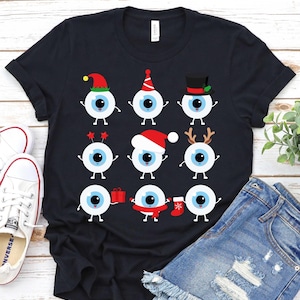 May include: Black t-shirt featuring nine cartoon eyeballs with festive holiday accessories. Each eyeball has a blue iris and white sclera, wearing hats, antlers, and holding gifts. The shirt is paired with denim shorts and white sneakers.