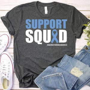 May include: A dark grey t-shirt with the words "SUPPORT SQUAD" in white with a blue ribbon in the middle of the word "SQUAD". The text "#DIGEORGESYNDROMEAWARENESS" is printed below the words.