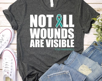 以下が含まれることがあります： 「NOT ALL WOUNDS ARE VISIBLE」の白文字と、ターコイズ色のリボン、そして「PTSD AWARENESS」がプリントされた濃いグレーのTシャツ。青いジーンズと白いスニーカーとコーディネートされています。