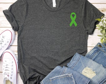 May include: A dark gray t-shirt with a green ribbon on the front. The t-shirt is laying on a white wooden surface with a pair of white sneakers and a pair of blue jeans.