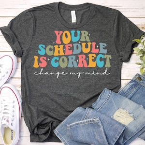 May include: A dark gray t-shirt with the text "YOUR SCHEDULE IS CORRECT change my mind" in colorful retro-style lettering.